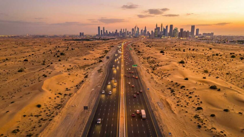 Aerial view of a multi-lane highway cutting through UAE desert landscape toward a distant city skyline at sunset